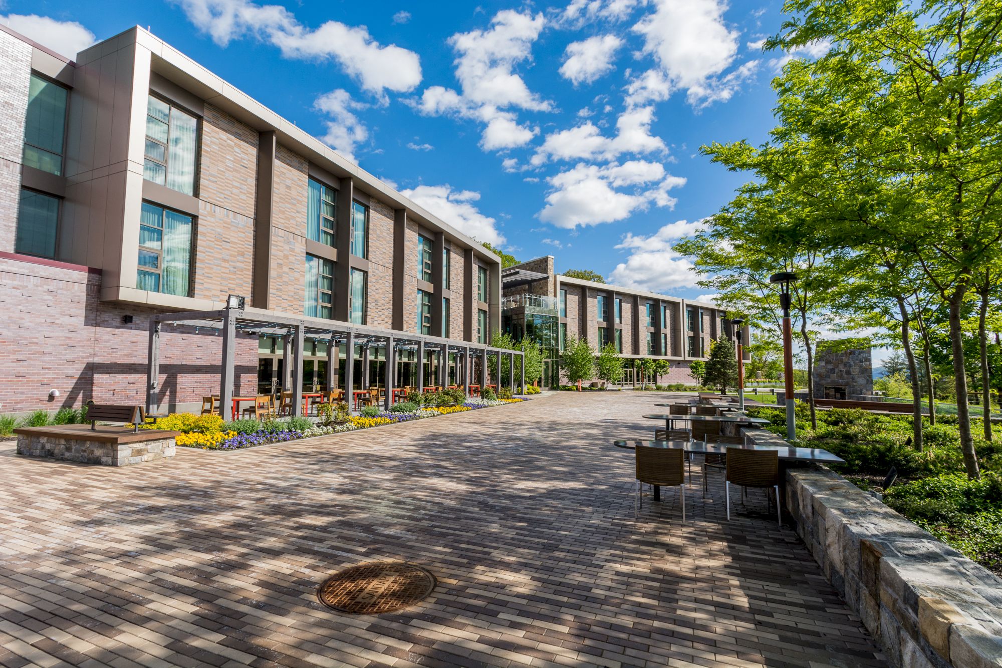 A modern building with large windows, a wide walkway, outdoor seating, vibrant flowers, and trees under a blue sky with clouds.